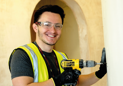 Male Construction Student Drilling In To A Wall And Wearing Hi Vis Jacket